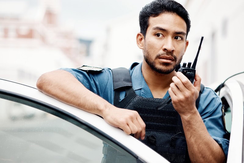 Asiatischer Mann, Polizei und Walkie Talkie für Funk in der Stadt Kommunikation, Verstärkung oder Notfall. Ernste männliche Person oder Wachmann im Polizeiauto und ruft Verstärkung für Verbrechen auf Patrouille in einer städtischen Stadt
