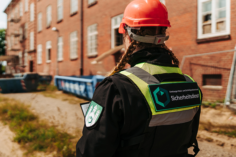 Eine Person mit Sicherheitsweste und rotem Helm steht vor einem Backsteingebäude.
