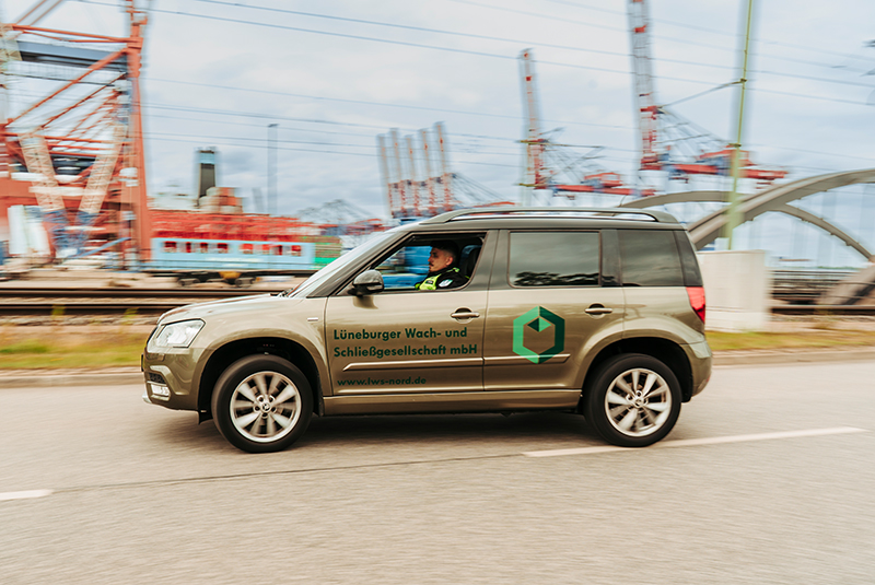 Ein grüner SUV mit einem Wach- und Sicherheitslogo fährt vor Industrierahmen.