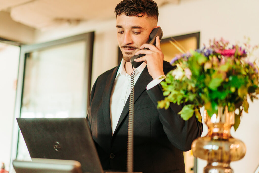 Ein junger Mann im Anzug telefoniert lächelnd am Schreibtisch mit Blumenstrauß